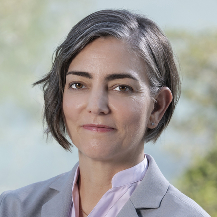 A woman with short, grayish hair wearing a light gray blazer and white shirt, outdoors with blurred greenery in the background.