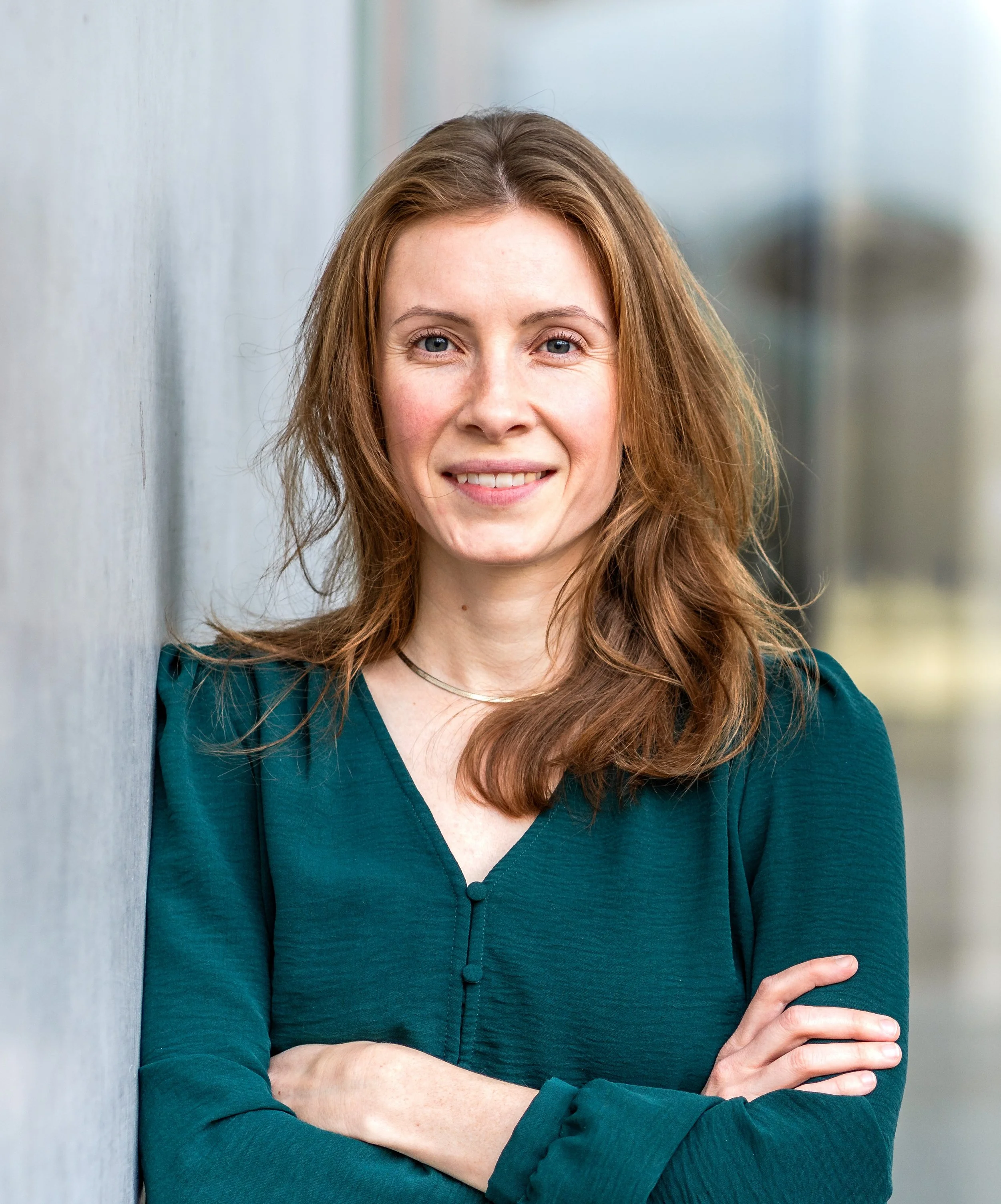 A smiling woman with long, wavy red hair standing outdoors, leaning against a gray wall with her arms crossed.