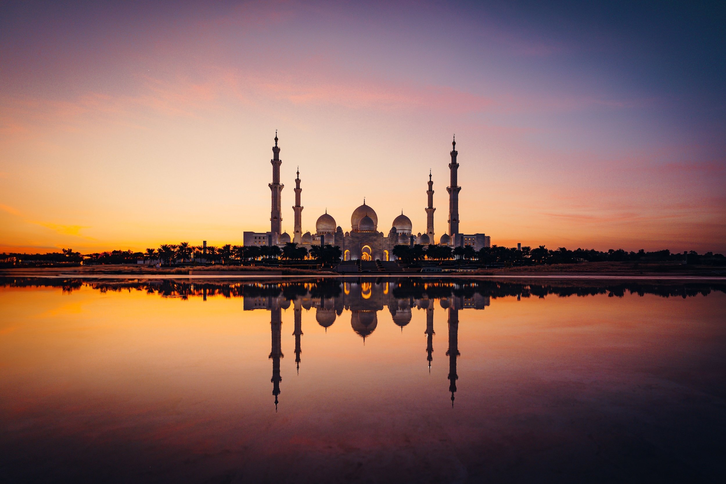 A mosque with multiple domes and tall minarets reflected in a body of water at sunset.