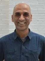 A man with a bald head and a broad smile, wearing a blue button-up shirt, standing in front of a white brick wall.