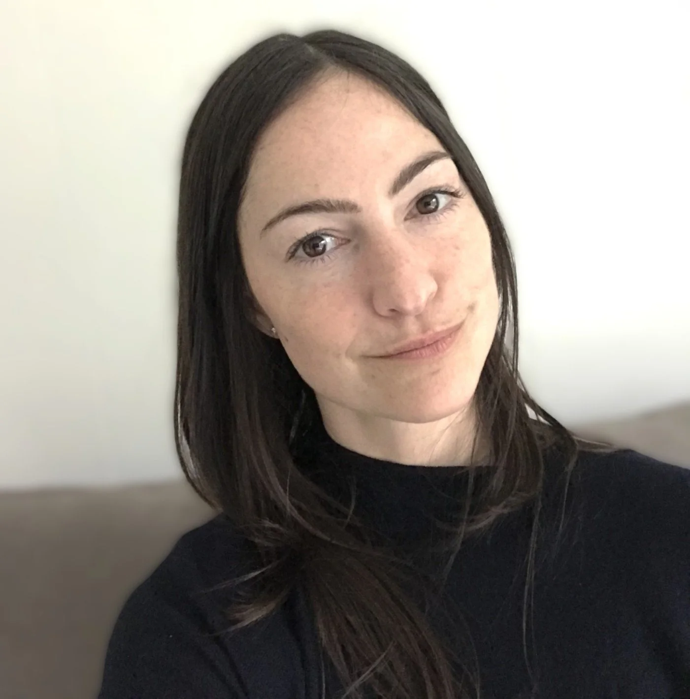 A woman with long dark hair, fair skin with freckles, and light-colored eyes, wearing a black top, sitting against a light-colored wall, smiling softly.