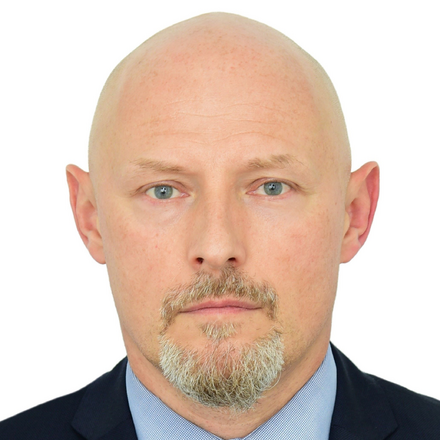 A bald man with a goatee and blue eyes wearing a dark suit and light blue shirt, looking directly at the camera against a white background.