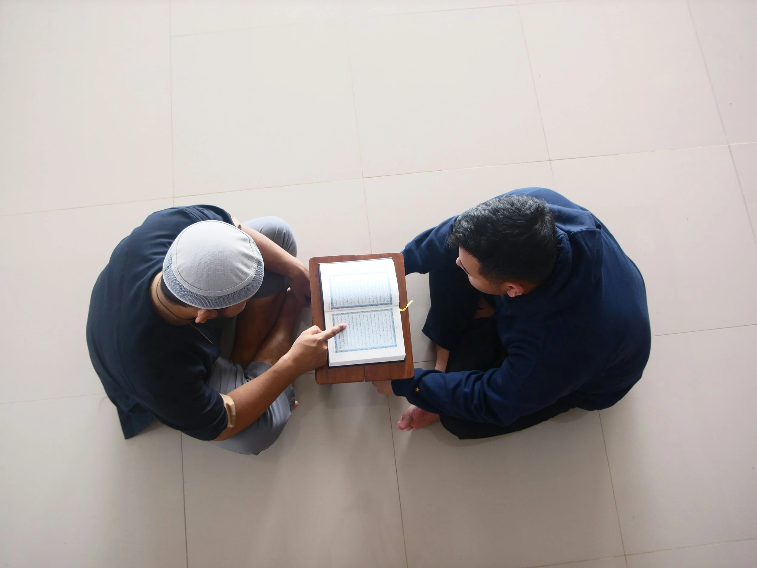 Two men sitting on the floor, one of them pointing at a book on a small table between them.