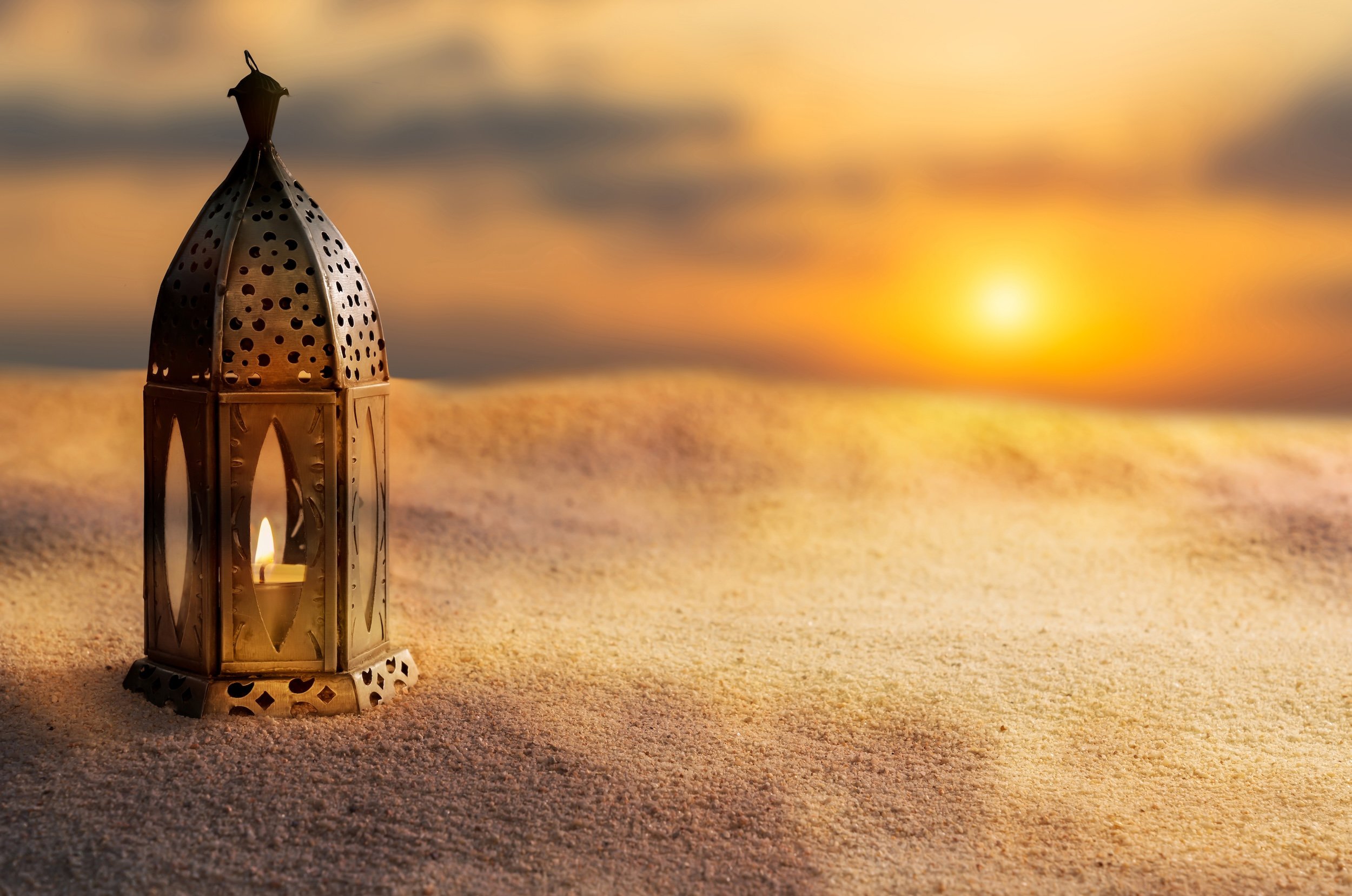 A decorative metal lantern with a lit candle inside, placed on sand at sunset with a blurred ocean and sky in background.