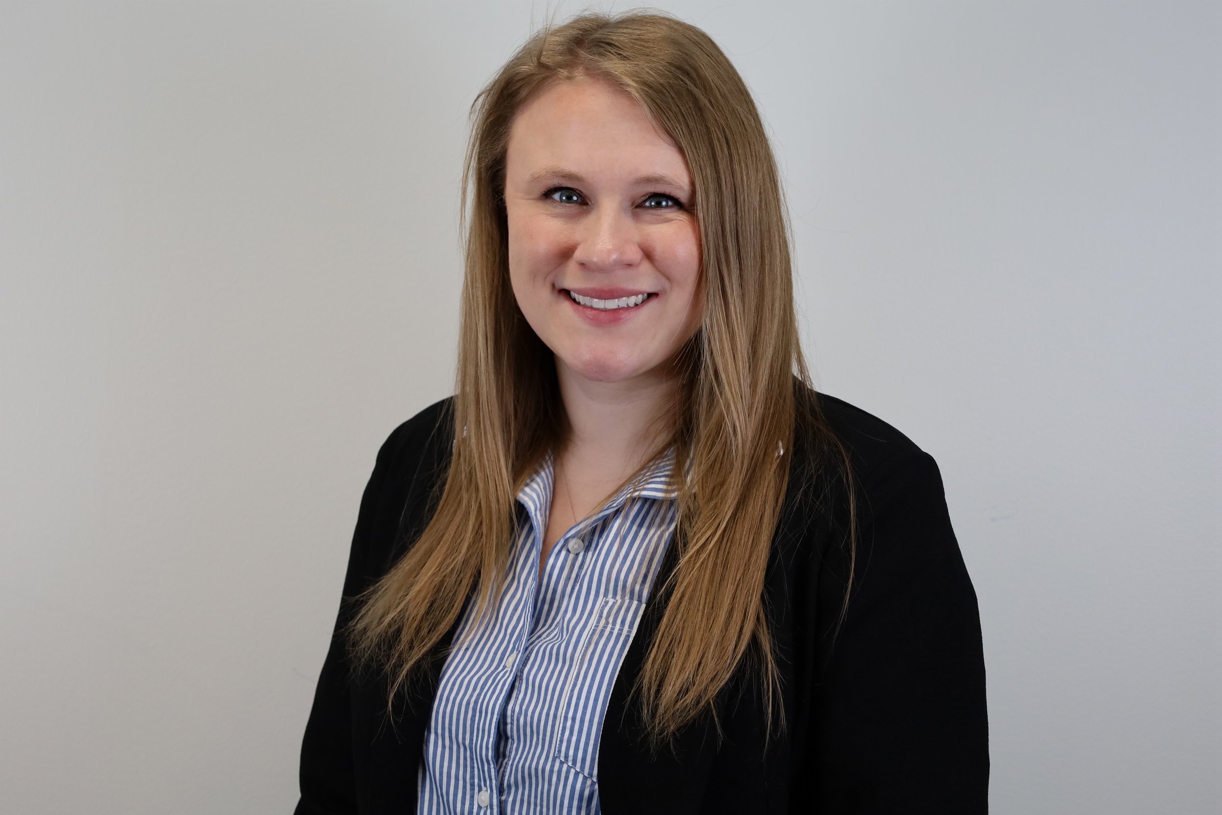 A woman with long red hair, blue eyes, and fair skin, smiling, wearing a black blazer over a blue and white striped shirt, standing against a plain light gray background.