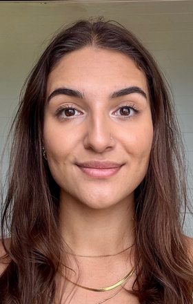 Close-up of a young woman with long brown hair, wearing a small nose hoop and a gold necklace, smiling softly at the camera.