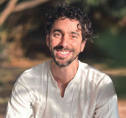 A man with curly dark hair and a beard smiling outdoors in natural light, wearing a light-colored shirt.