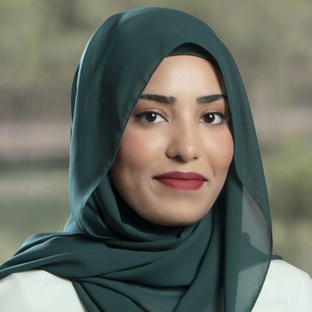 A woman wearing a dark green hijab with a confident smile indoors with a blurred window background.