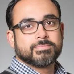 Close-up of a man with dark hair, glasses, and a beard, wearing a checkered shirt and dark jacket, against a neutral background.