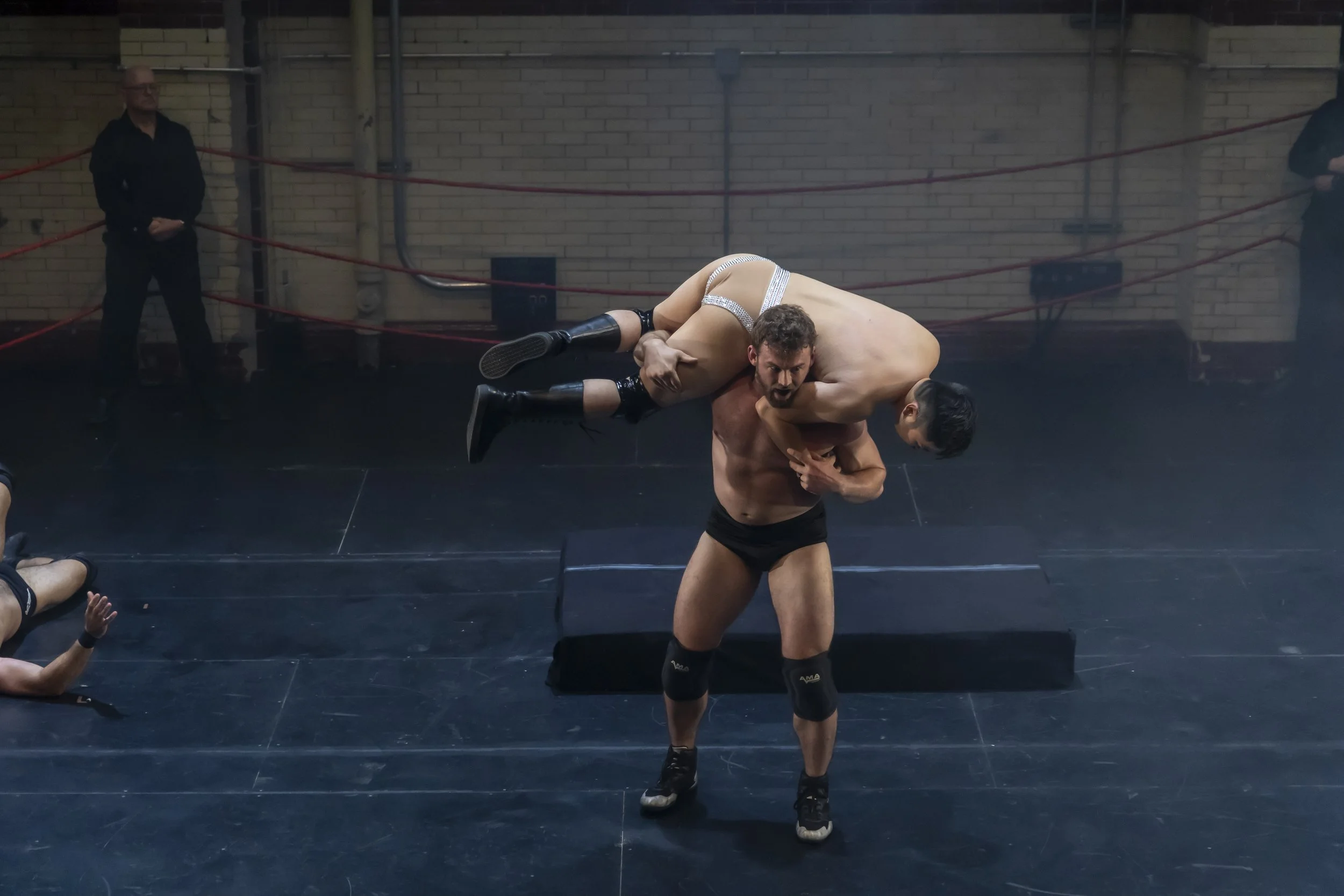 Two male wrestlers in a wrestling ring, with one lifting the other. The wrestler being lifted is barefoot and dressed in wrestling attire, while the other is wearing shoes and knee pads. There are officials or referees on the sides and a brick wall b