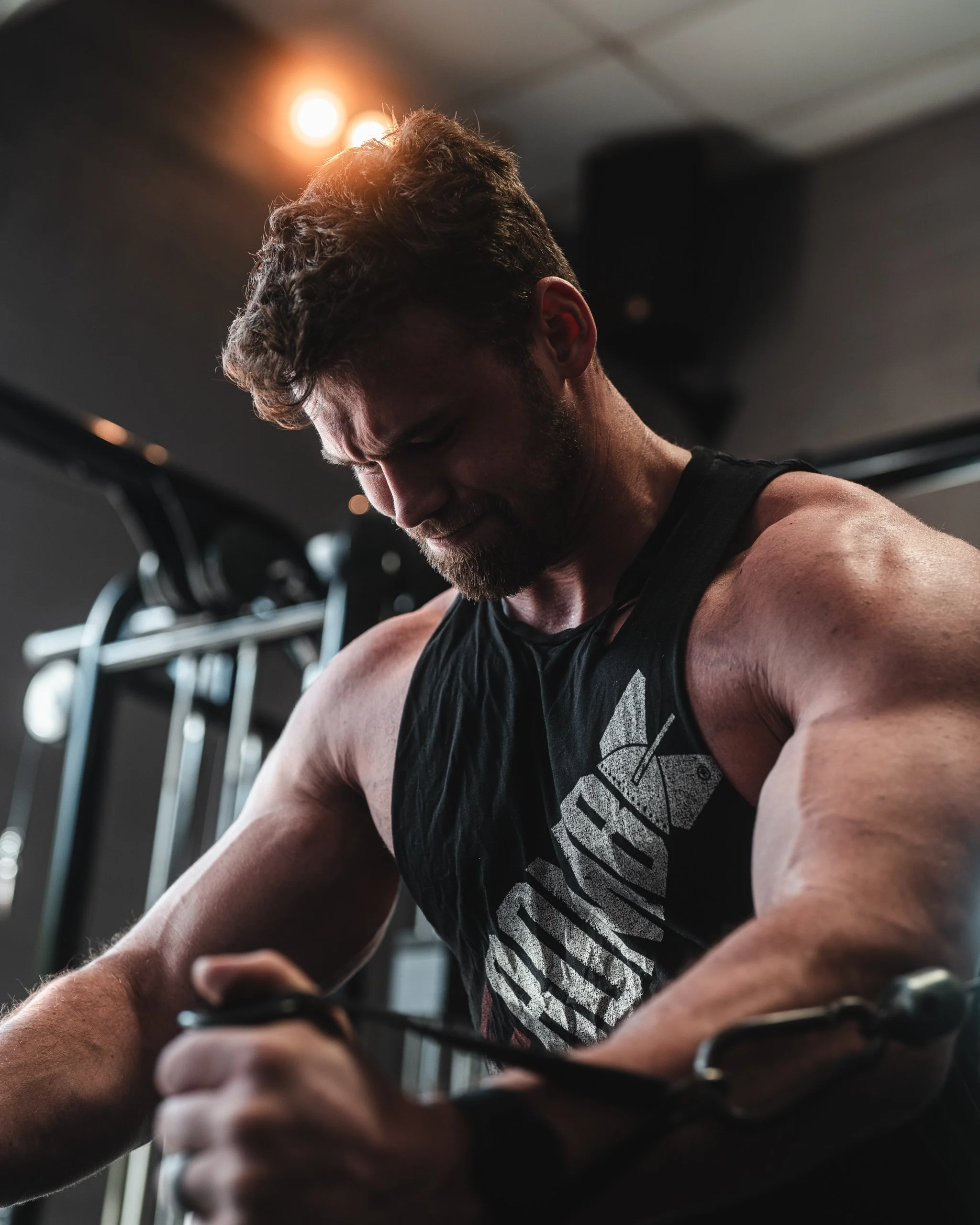 A muscular man with a beard and short hair working out at the gym, focusing on his arm muscles while holding a gym equipment.