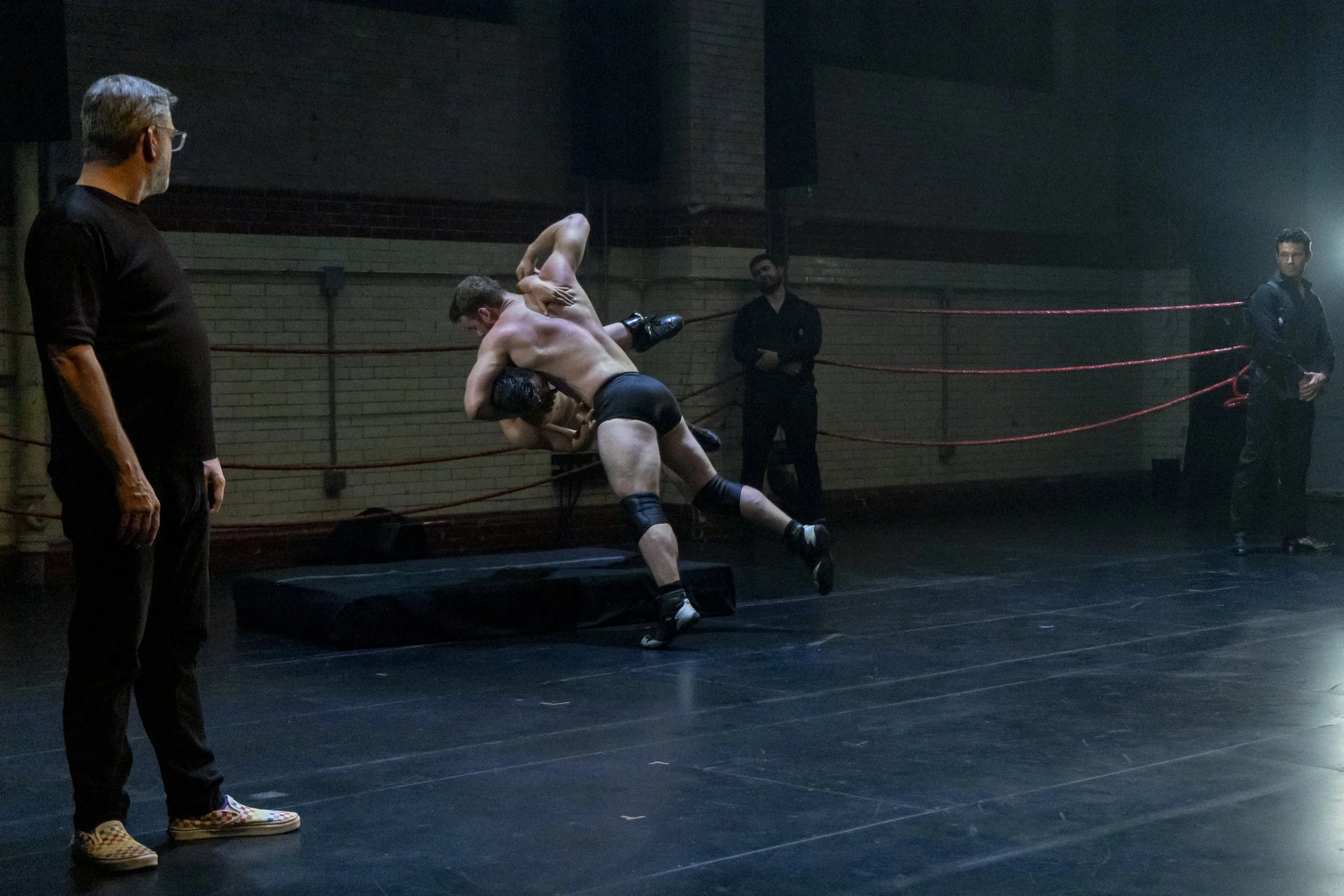 Two male wrestlers engaged in a wrestling match inside a ring with red ropes, with three spectators observing and a man in black standing nearby in a dimly lit room.
