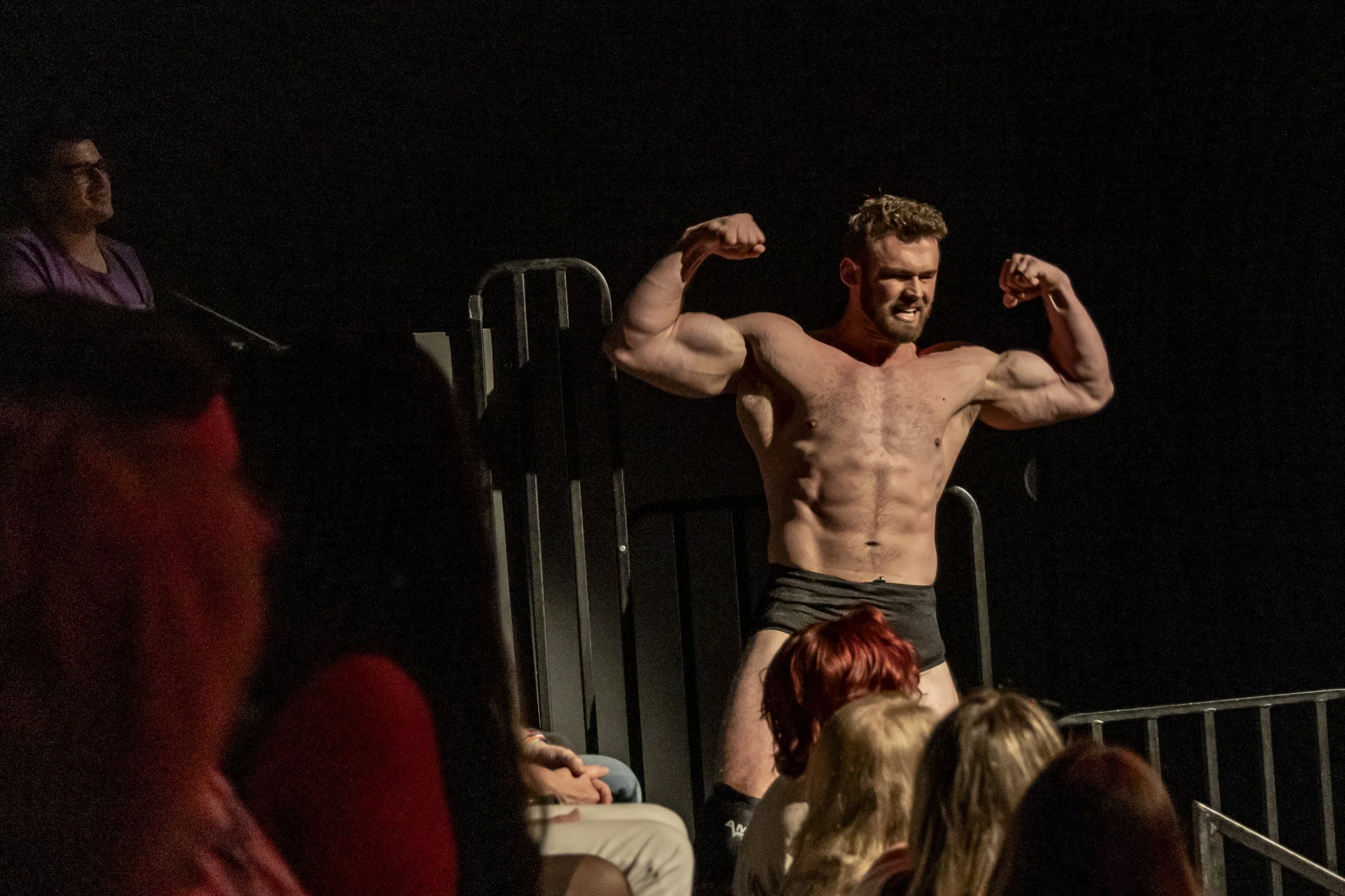 A muscular man posing on stage with arms flexed, wearing only black shorts, in front of a seated audience with some red-haired individuals, a woman with glasses and dark hair, and a person with bright red hair, in a theatrical or performance setting.