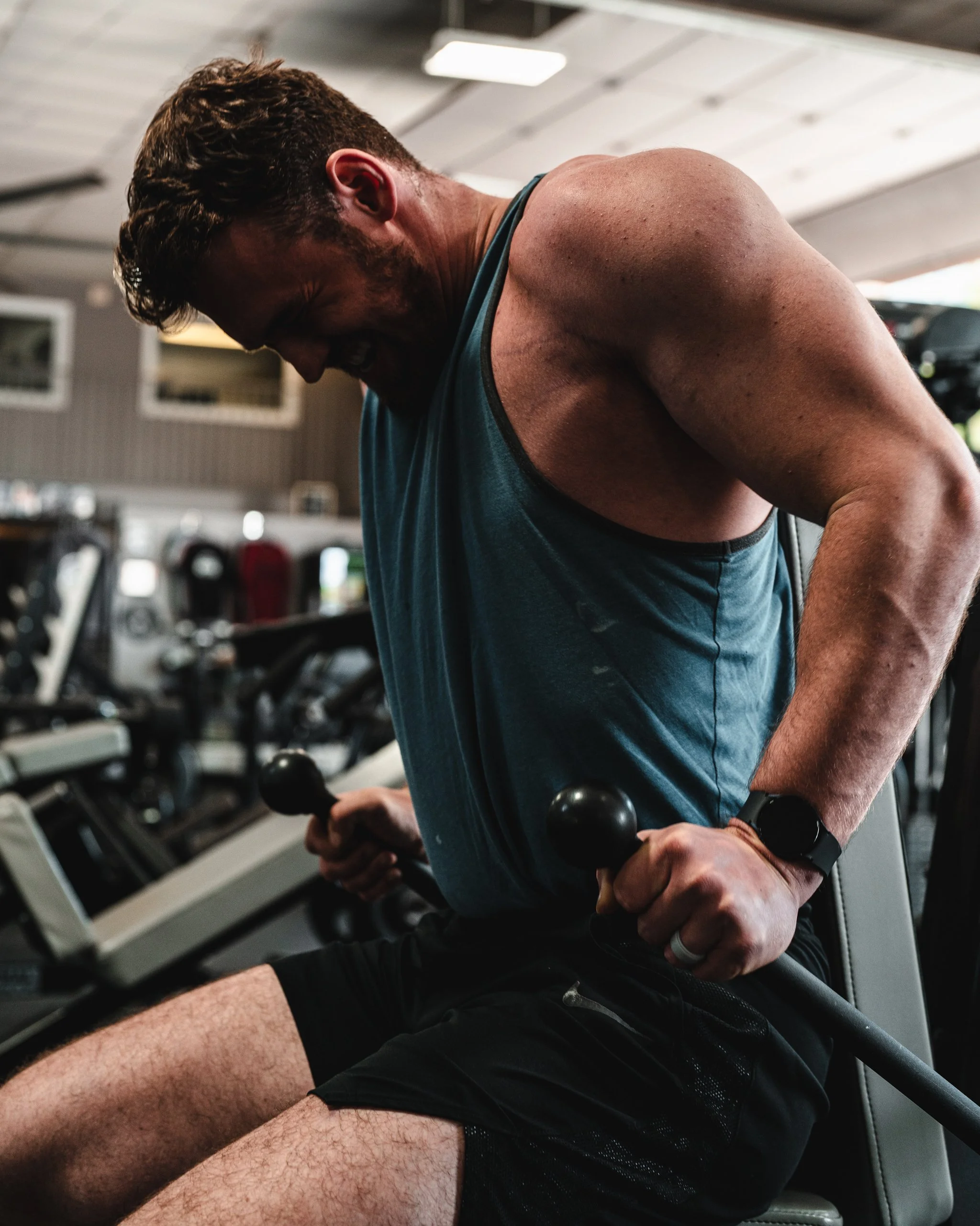 A man in a gym performing a tricep exercise with a dip bar, smiling and wearing a blue tank top, black shorts, and a fitness tracker on his wrist.