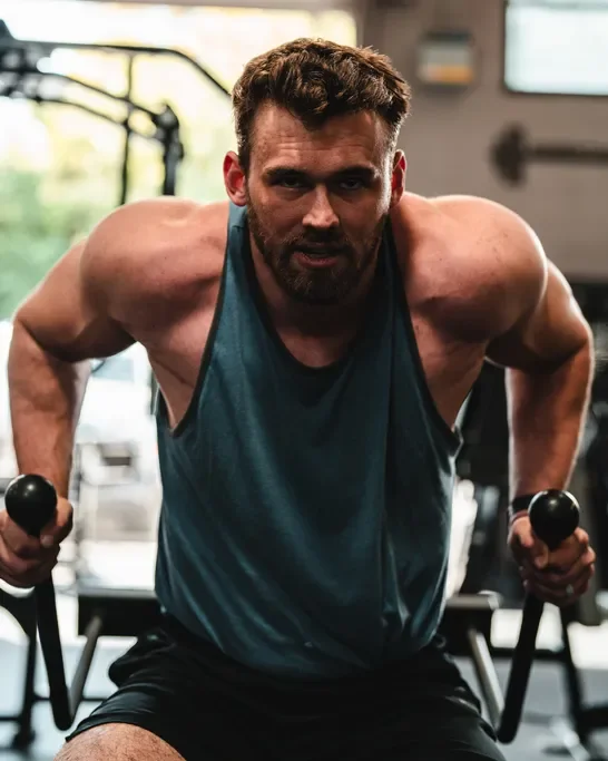 A man with a muscular build and beard working out with dumbbells in a gym.