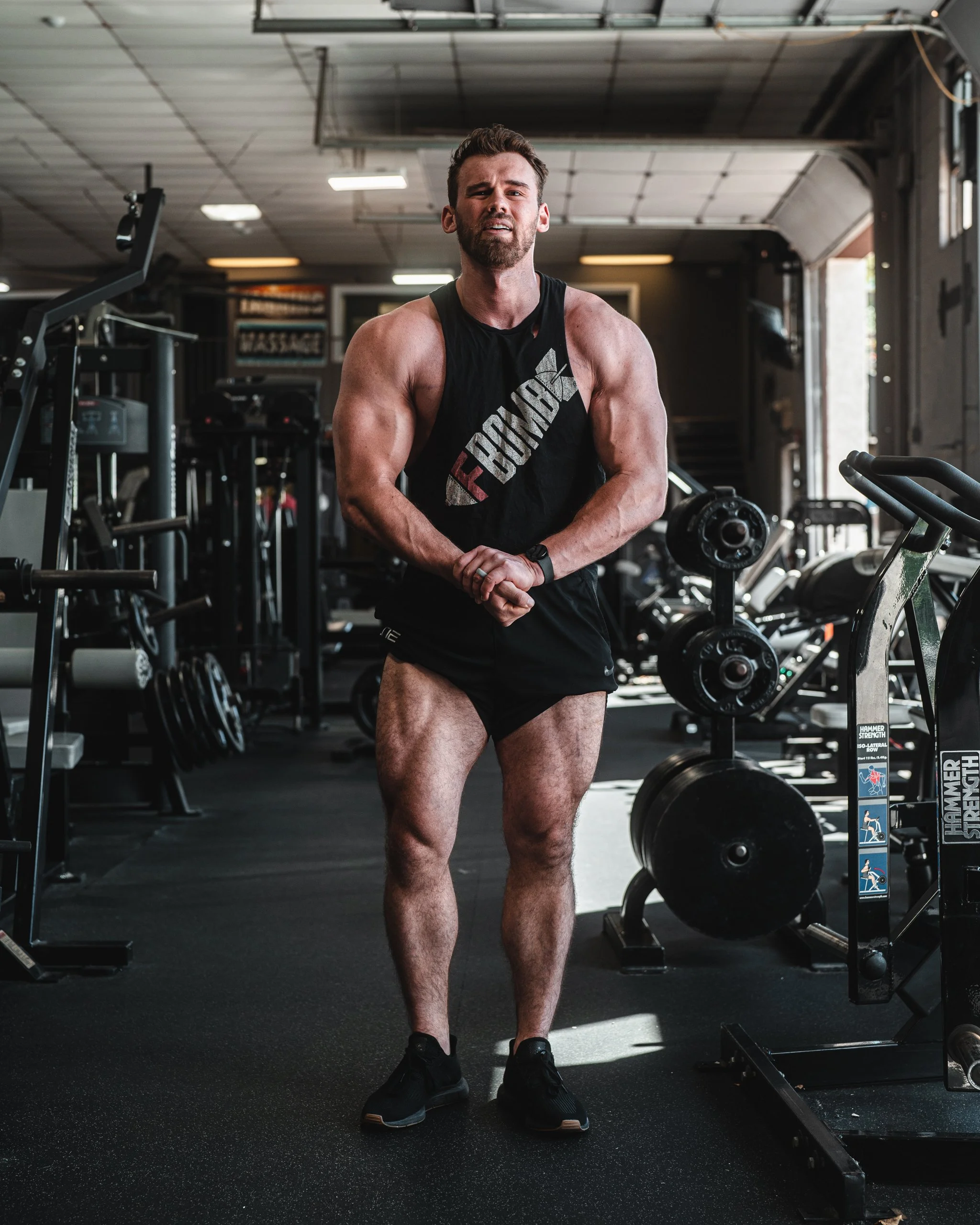 A muscular man with a beard standing in a gym, wearing a black sleeveless workout shirt and black shorts, with gym equipment around him.