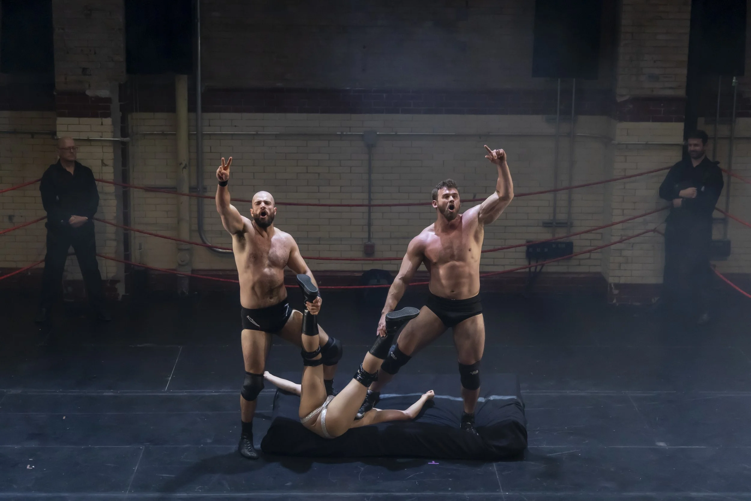 Two shirtless wrestlers standing inside a wrestling ring with red ropes, one holding a microphone, and a woman in professional wrestling attire lying on the mat between them, while two security guards stand outside the ring watching.