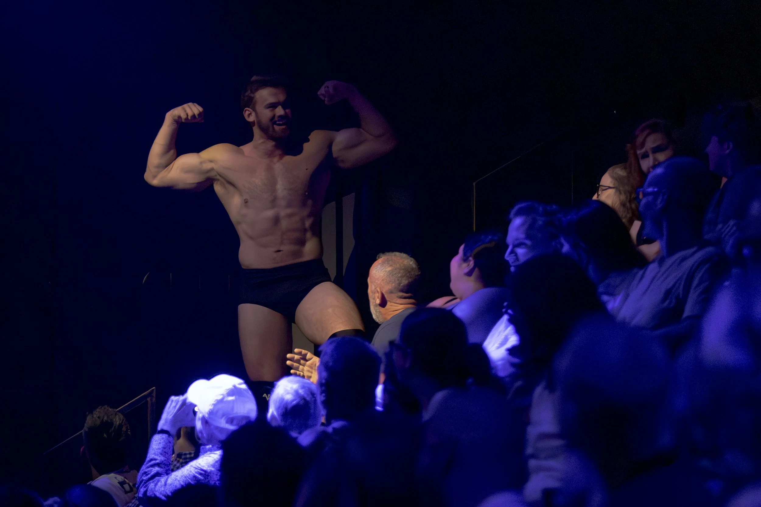A shirtless man with a muscular build and dark hair is posing confidently in front of an audience, wearing black underwear, in a dimly lit setting.