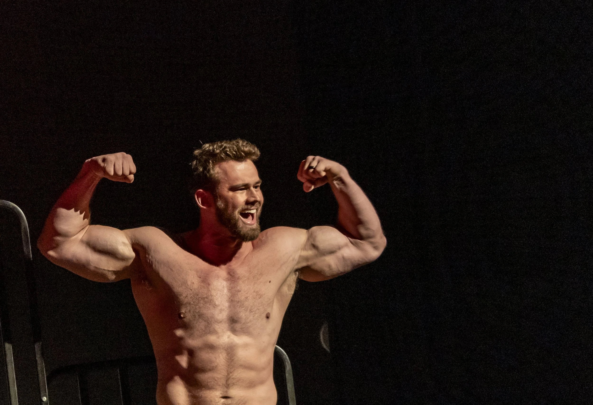 A shirtless man with a beard and curly hair flexing his biceps and smiling against a dark background.