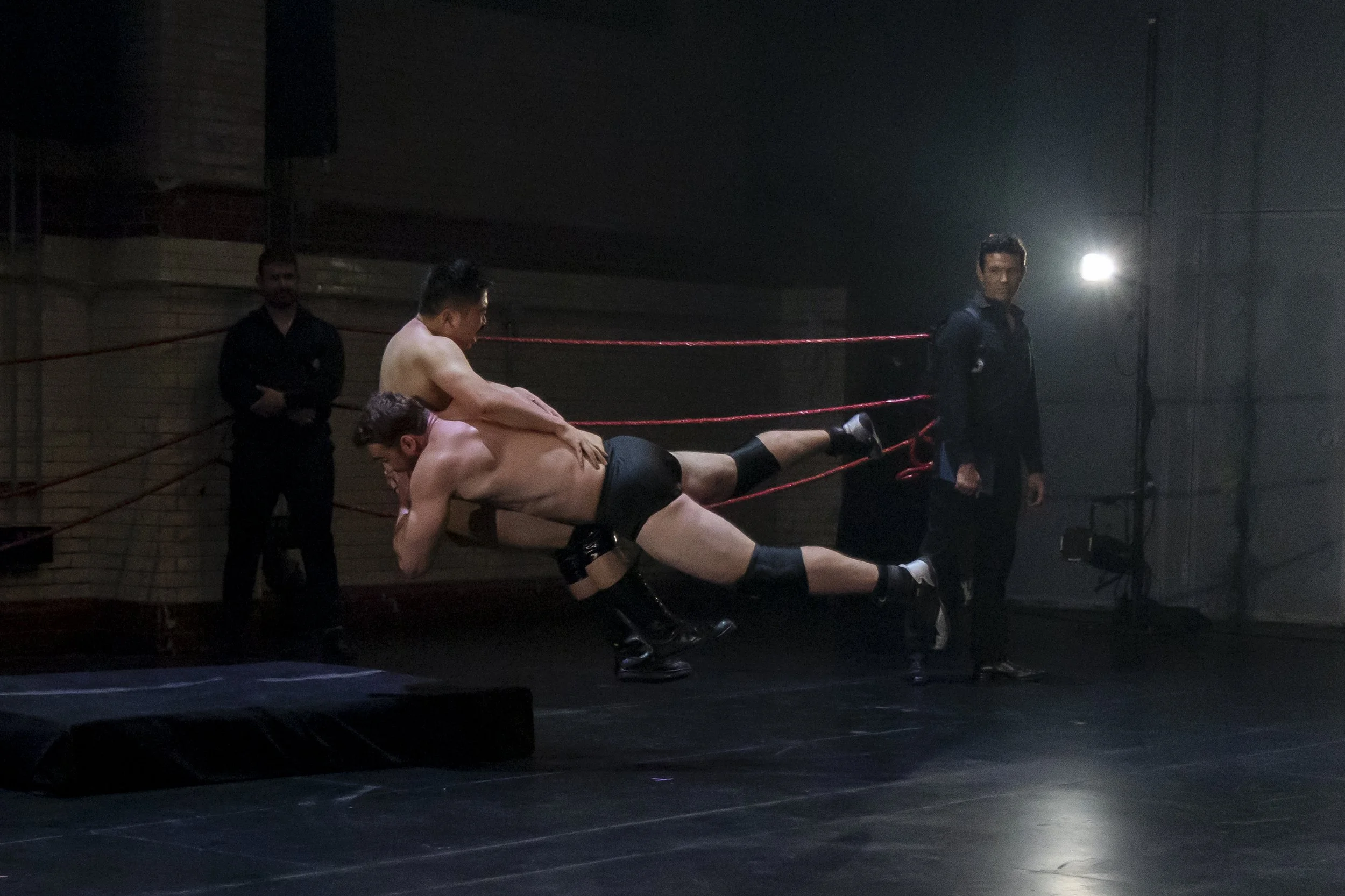 Two men wrestling in a wrestling ring with ropes, one is lifting the other, while two other men in dark clothing watch in a dimly lit room.