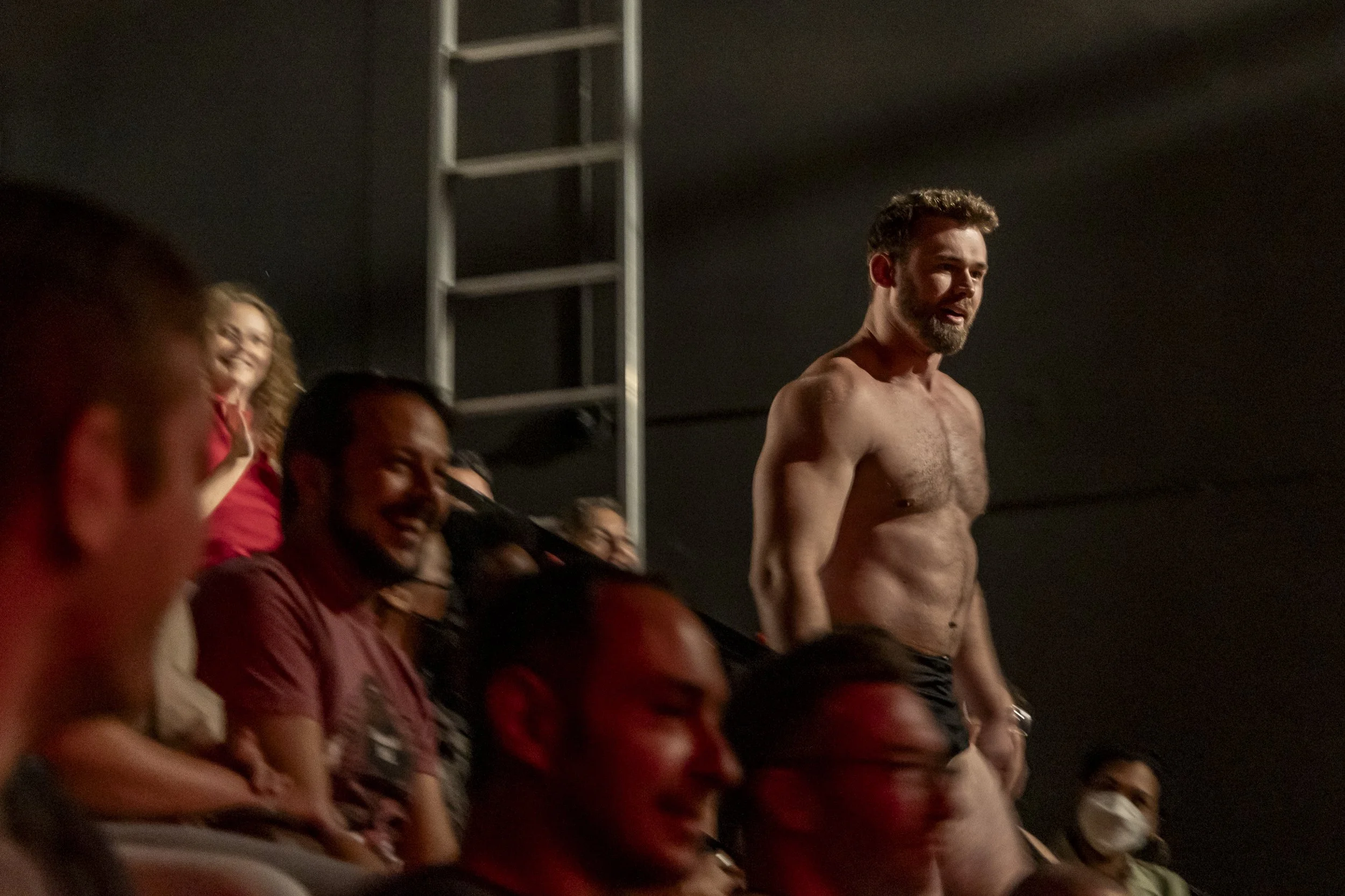 A shirtless man with a beard standing in front of an audience, smiling, while people in the audience look at him and smile or laugh, some wearing face masks.