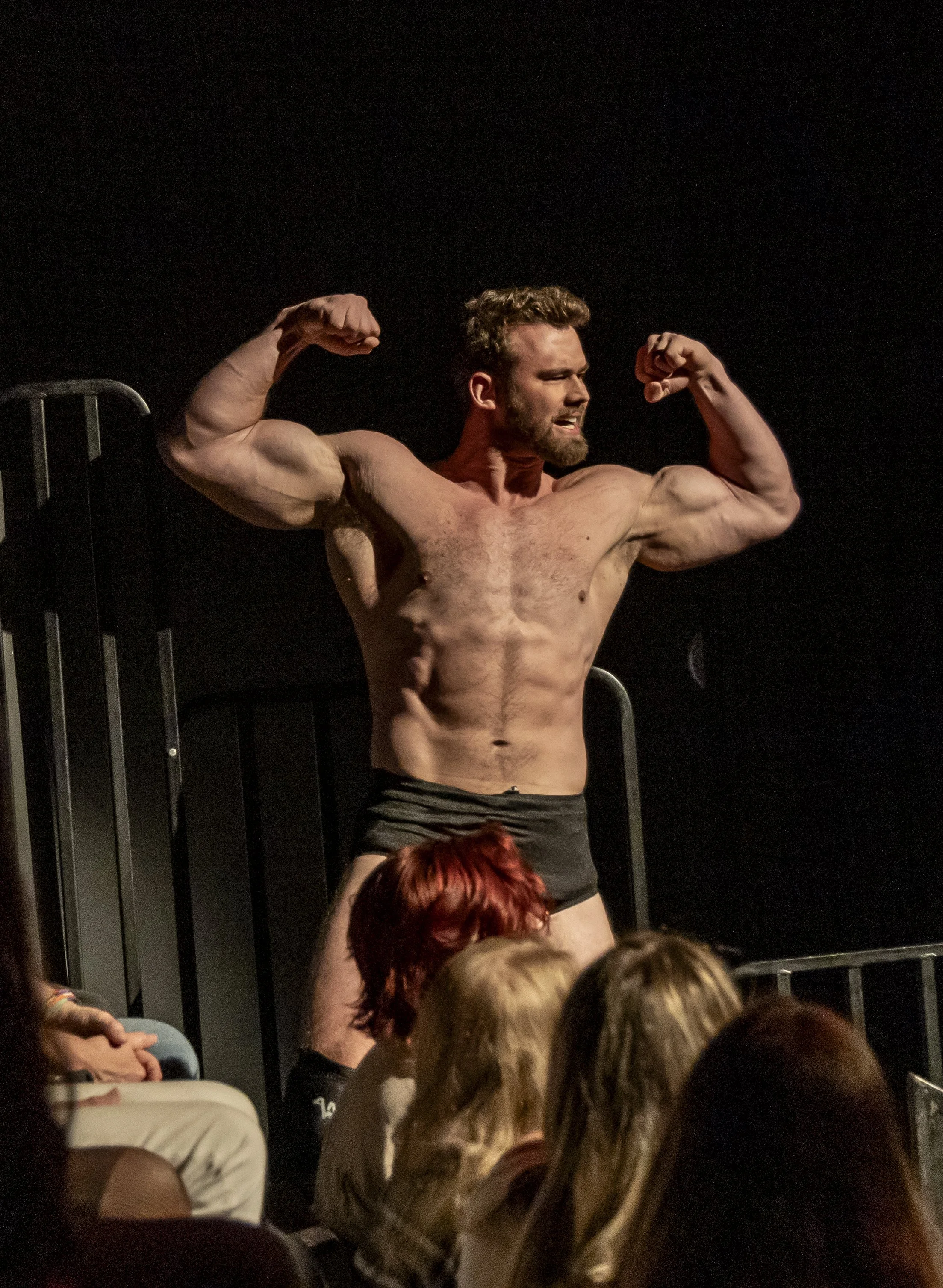 A muscular man with short hair and a beard, wearing black shorts, is flexing his biceps on stage in front of an audience. The background is dark, and there are people seated in front of him, watching.