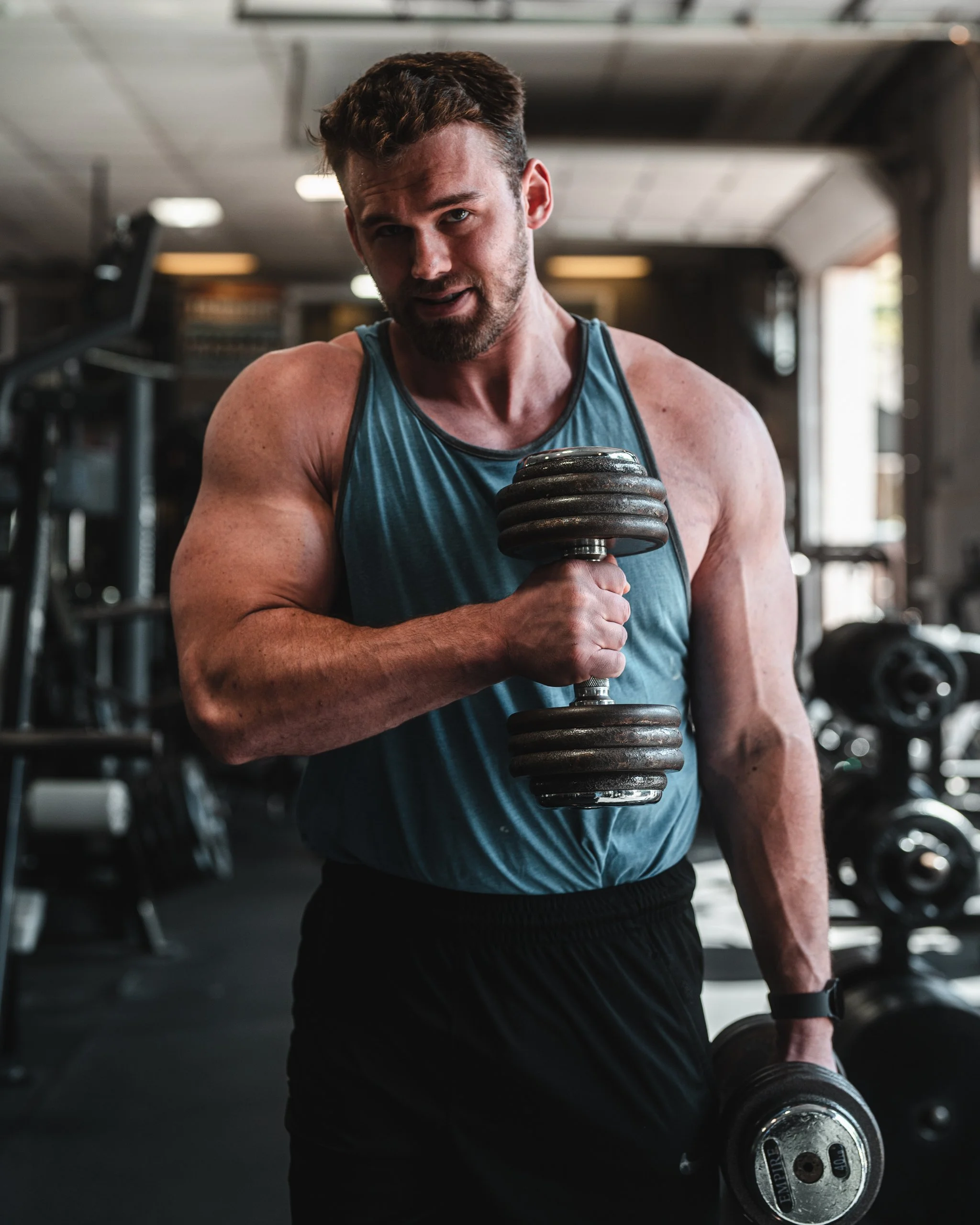 A muscular man in a gym, wearing a blue sleeveless shirt, holding a dumbbell in one hand, and looking at the camera.