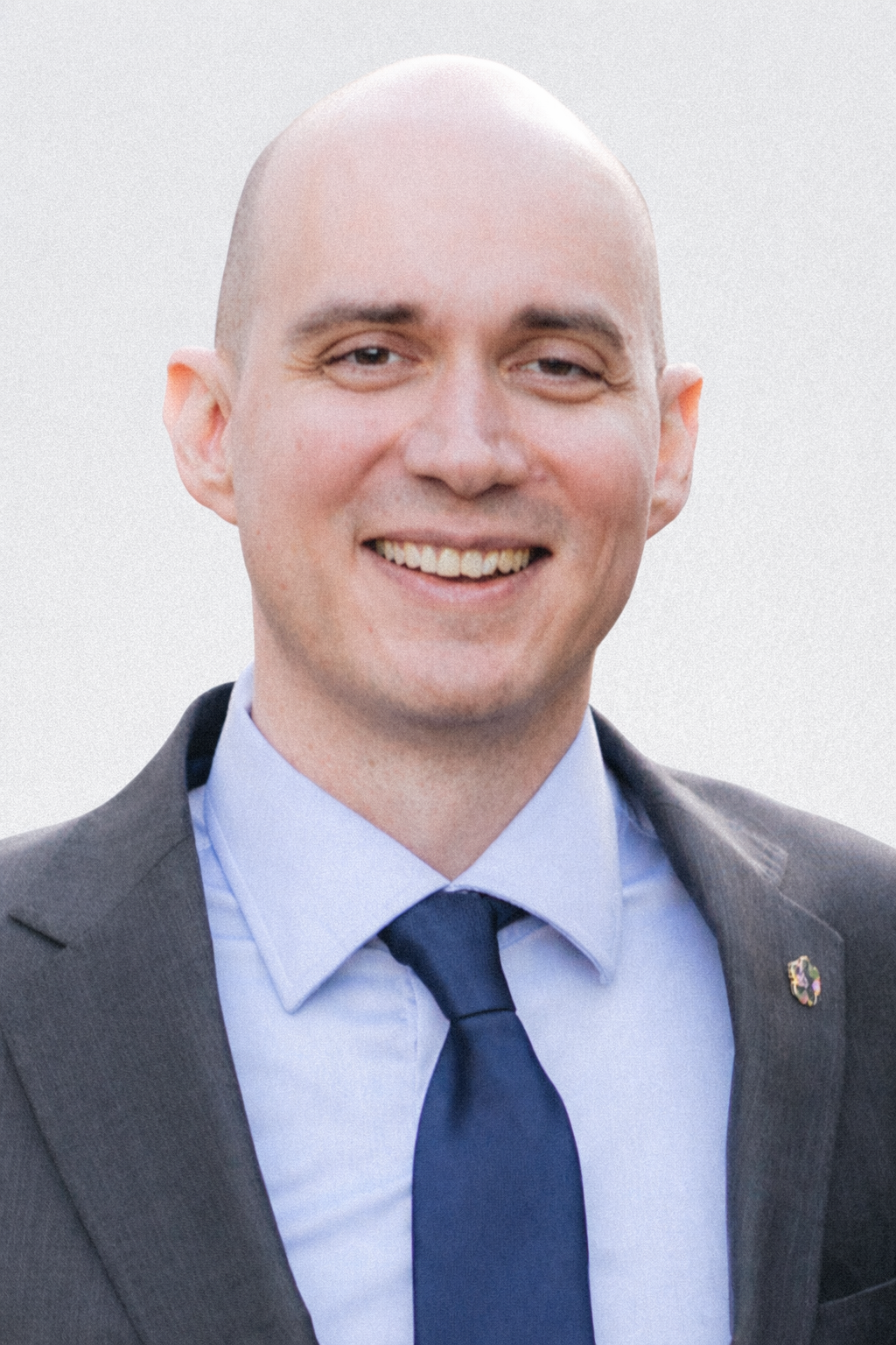 A smiling man in a suit and tie, with a light blue shirt, in an outdoor setting.