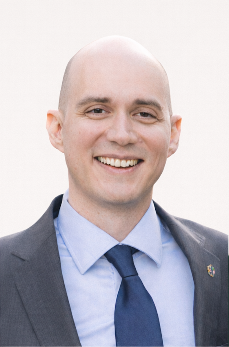 A smiling man in a gray suit, light blue dress shirt, and navy tie against a plain light background.