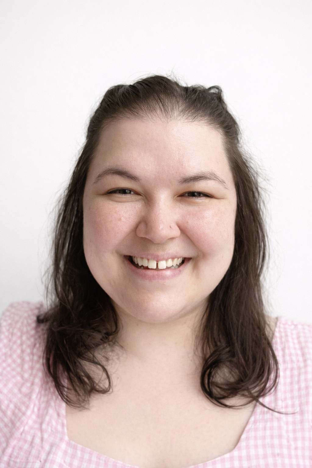 A young woman smiling with dark brown wavy hair wearing a pink and white checkered top.
