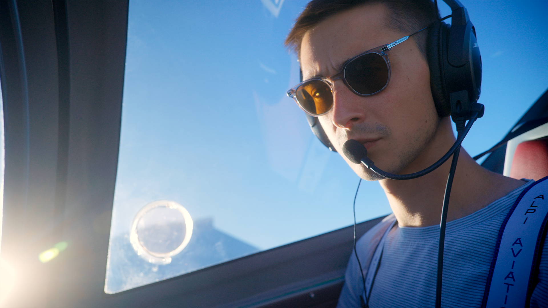 Gros plan de Raphaël dans le cockpit de l'avion en plein vol, lunettes de soleil et casque sur les oreilles. Ciel bleu en arrière plan et soleil qui fait un flare sur l'image.