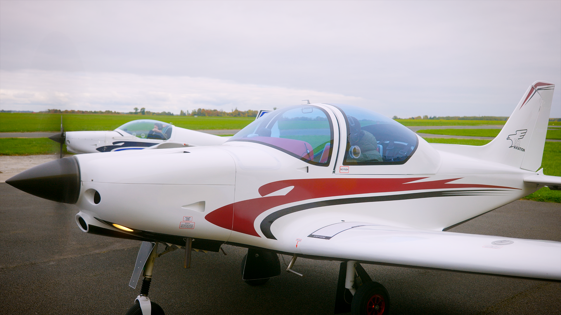 Deux avions légers de coloris blanc avec des accents rouges et noirs, stationnés sur un tarmac, avec un champ vert et un ciel nuageux en arrière-plan.