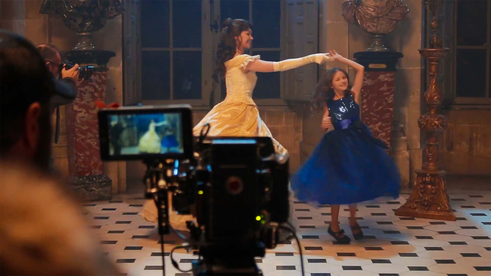 La Princesse Belle dans sa robe jaune et une petite fille en robe bleue foncée dansent dans une salle de château décorée avec des colonnes en marbre. Elles sont filmées par une caméra au premier plan.