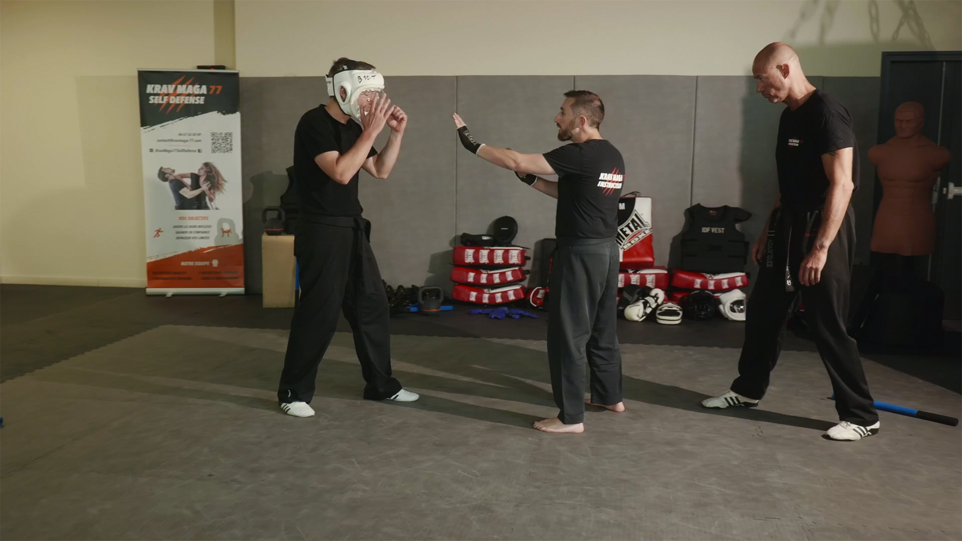 Trois hommes dans une salle de sport, un avec un casque de protection, s'entraînent au krav maga.