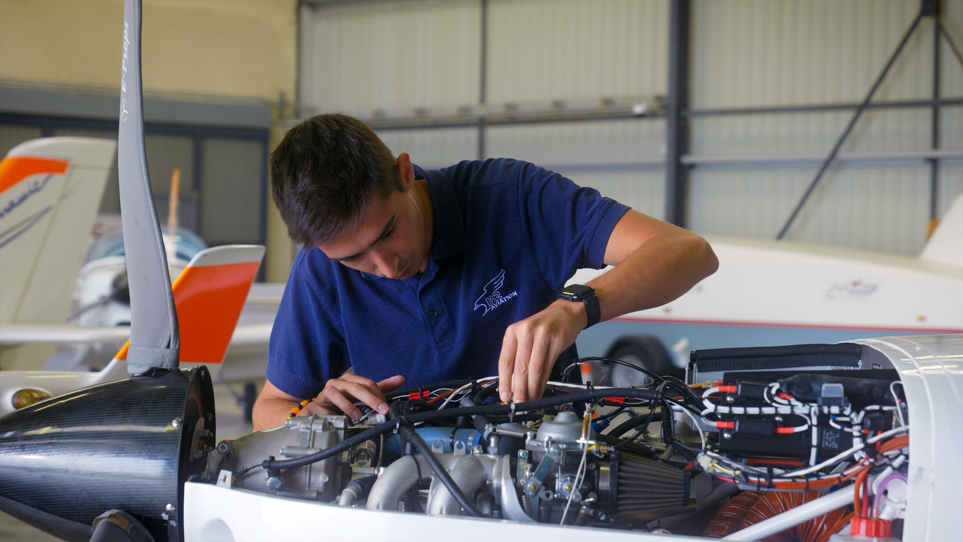 Un jeune homme en polo bleu travaille sur un moteur d'avion dans un hangar, entouré de petits avions.