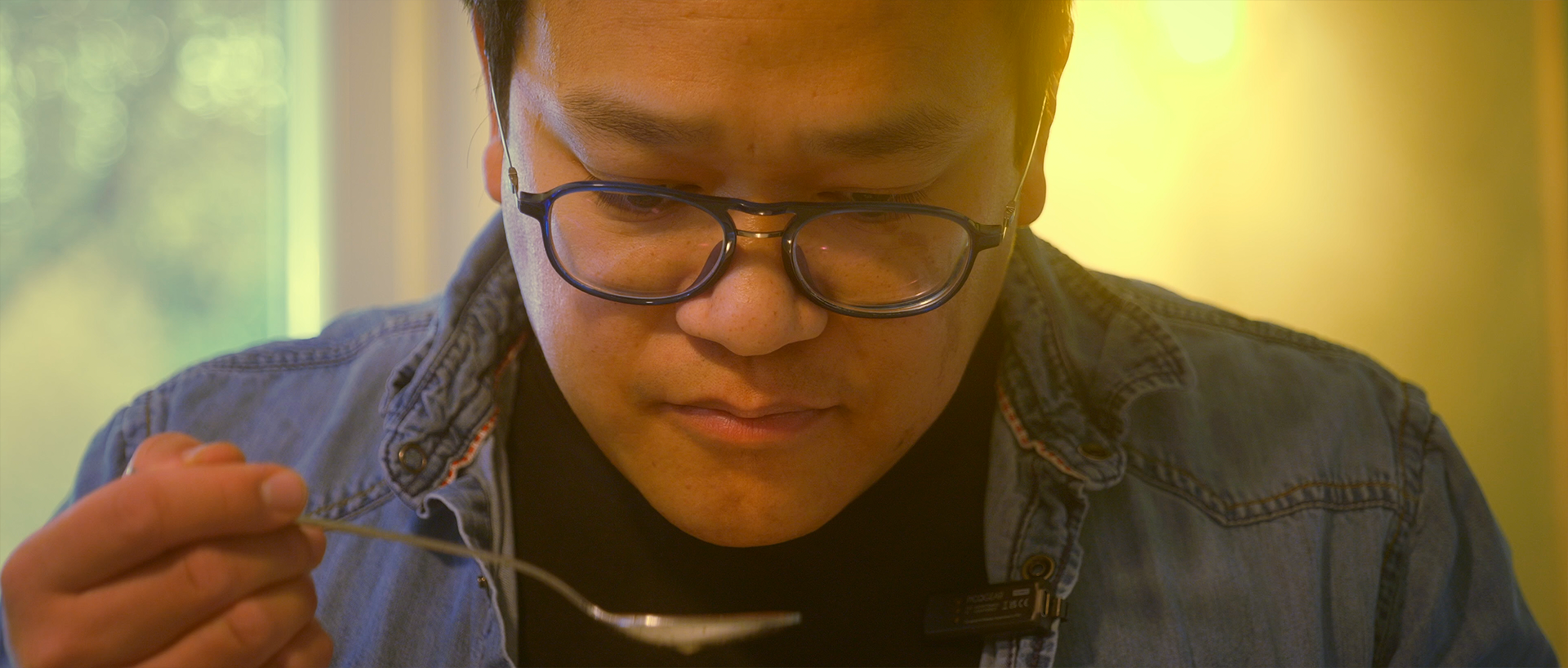 Gros plan de Philippe, homme asiatique portant des lunettes, tête baissée en goûtant un plat avec une cuillère, une lumière douce en arrière-plan.