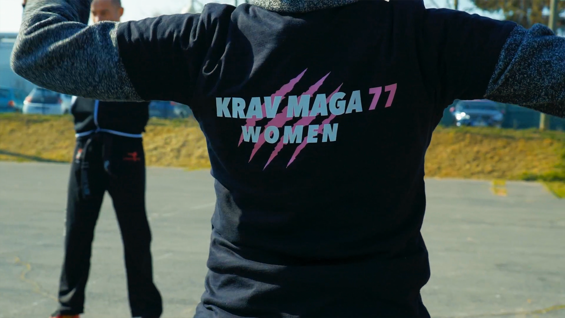 Gros plan d'une personne portant un t-shirt noir avec le texte 'KRAV MAGA 77 WOMEN' en violet clair et des griffures roses, en pleine séance d'entraînement en extérieur.
