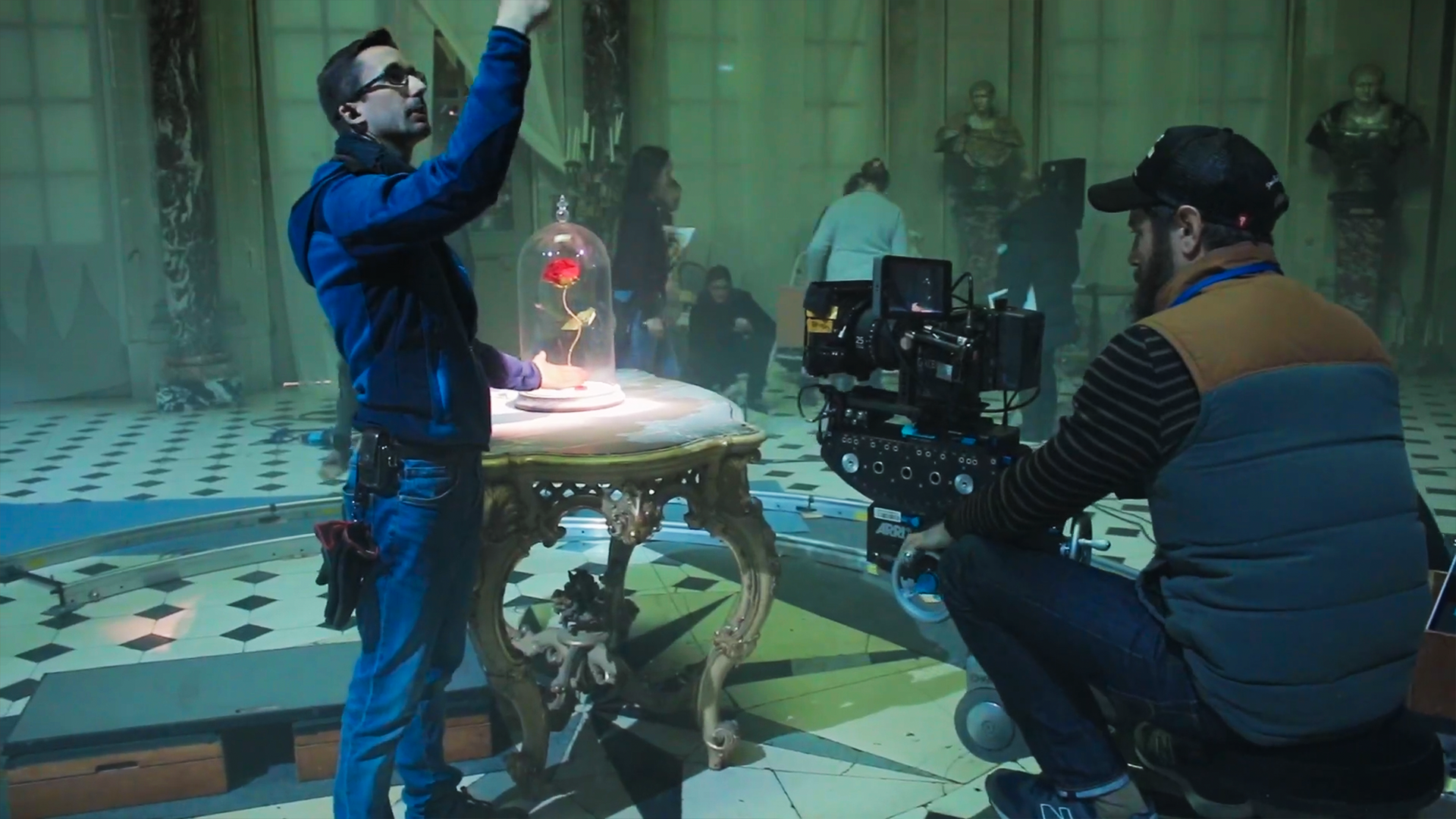 Equipe de tournage qui prépare une scène avec un homme debout qui règle une lumière et un autre assis derrière la caméra. Ils filment une rose sous une cloche sur une table. des personnes qui travaillent et des statues antiques.