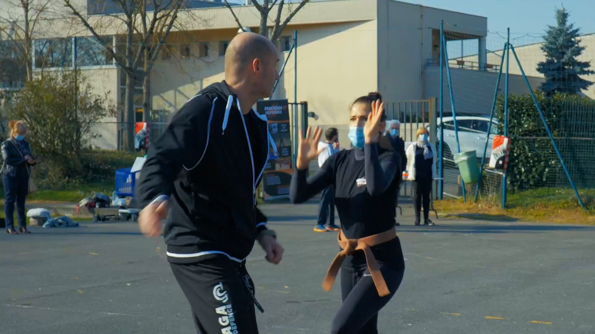 Un homme et une femme en tenue de sport font une démonstration de self-défense dans une cour extérieure, avec d'autres personnes en arrière-plan.