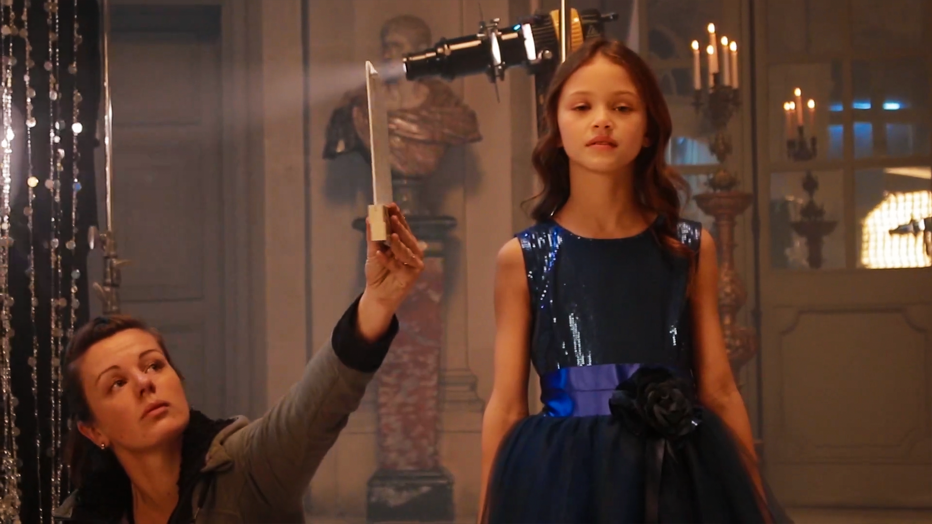 Une fillette en robe bleue, une femme technicienne tenant un clap juste à ses côtés. Devant un décor avec des effets de lumières et des chandeliers.