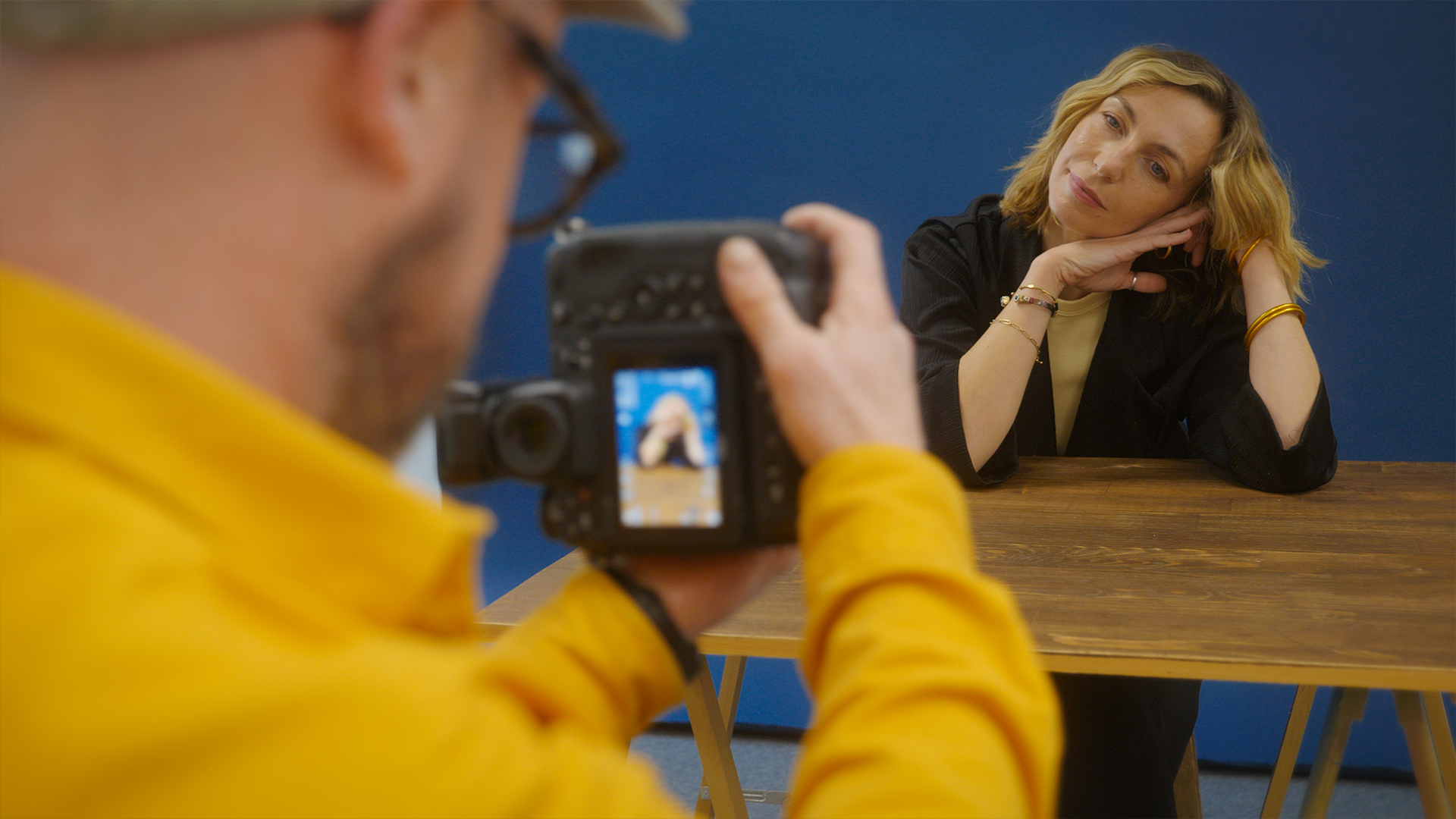 Une femme blonde pose pour une séance photo devant un fond bleu, assise à une table en bois. Manuel, chemise jaune, en amorce au premier plan, avec son appareil, la photographie.