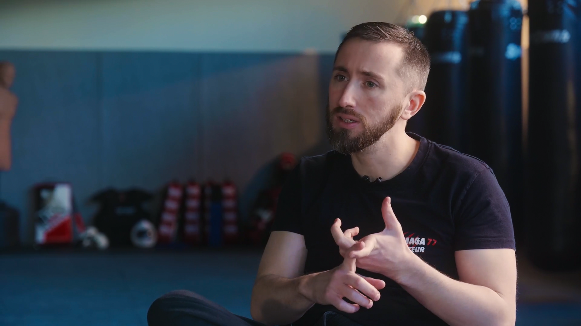 Guillaume, jeune homme avec une barbe et une coupe de cheveux courte parle en interview, assis dans un dojo, avec des sacs de boxe et des équipements sportifs visibles en arrière-plan.