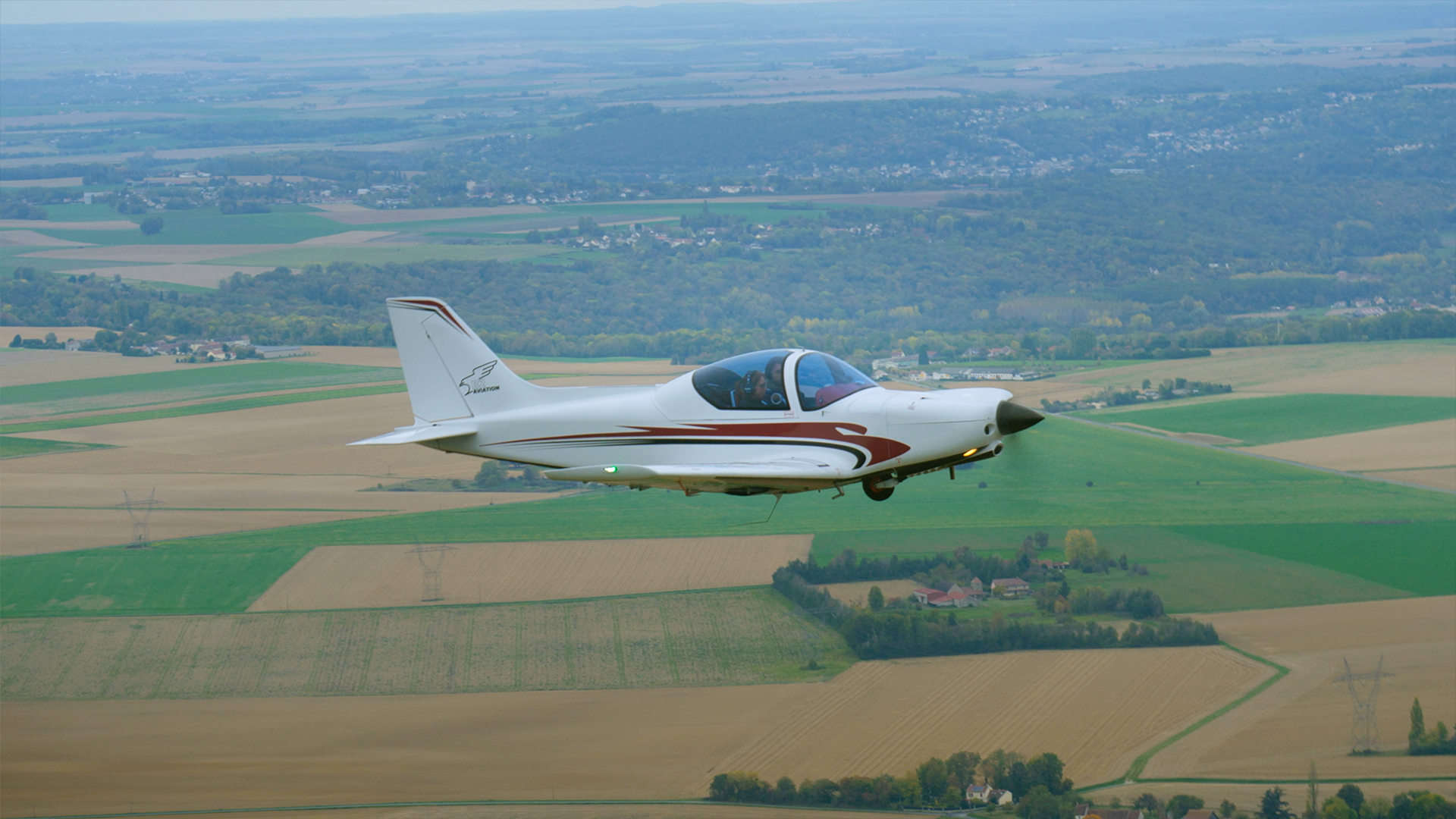 Un avion léger blanc avec des lignes décoratives rouges volant au-dessus d'une campagne verte et parsemée de champs agricoles.