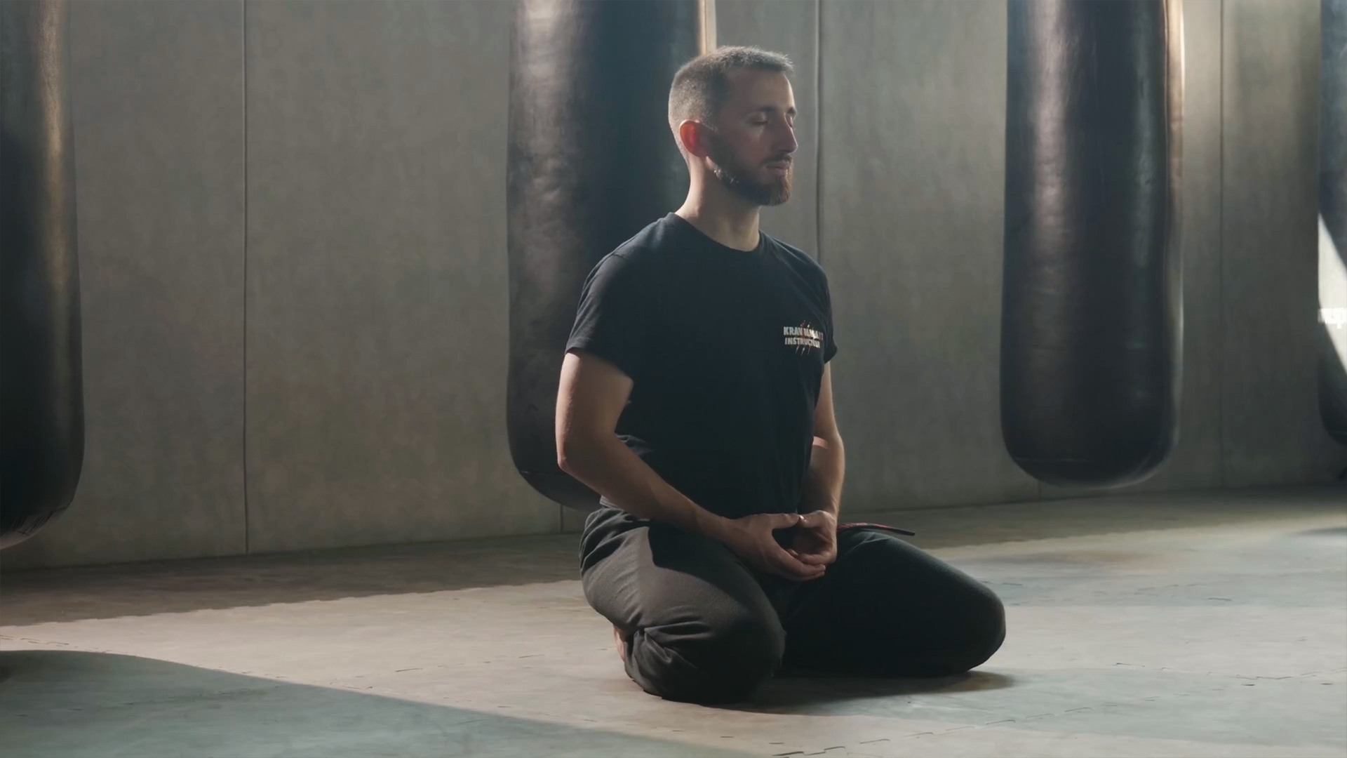 Guillaume, à genoux dans une salle de sport devant des sacs de frappe, médite, baignant dans une lumière douce du soleil.