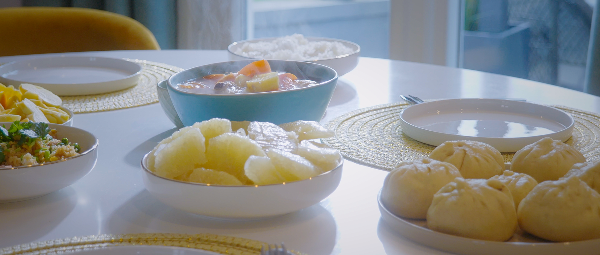 Table de repas asiatique avec des beignets, une salade, des fruits cuits, une soupe, et des bols de céréales, illuminée par les rayons du soleil.