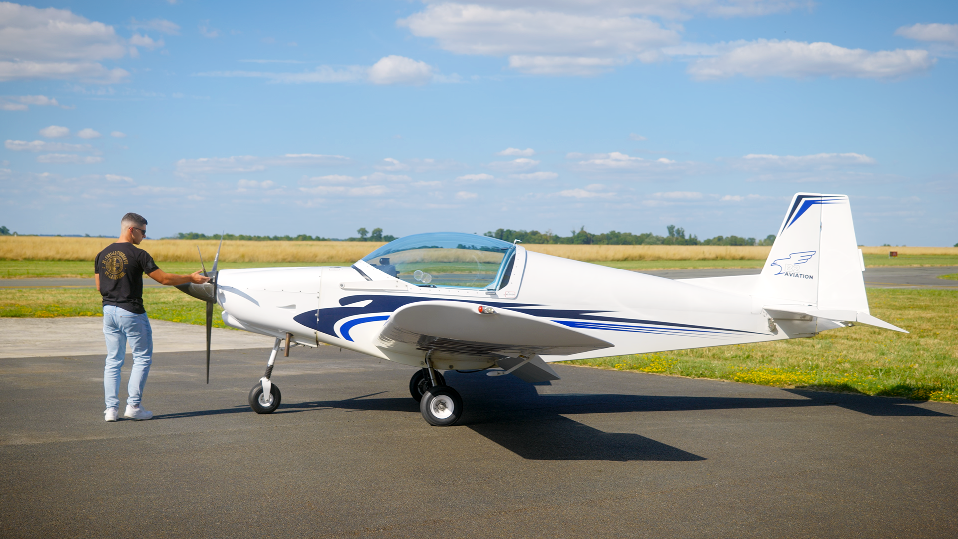 Un homme se tient près d'un petit avion blanc et bleu sur une piste d'aérodrome, préparant l'avion pour le vol.
