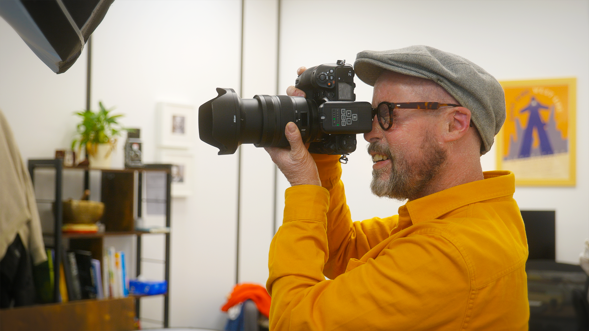 Manuel vu de profile, avec sa casquette, ses lunettes et sa chemise jaune, prend une photo avec un appareil professionnel dans son studio.