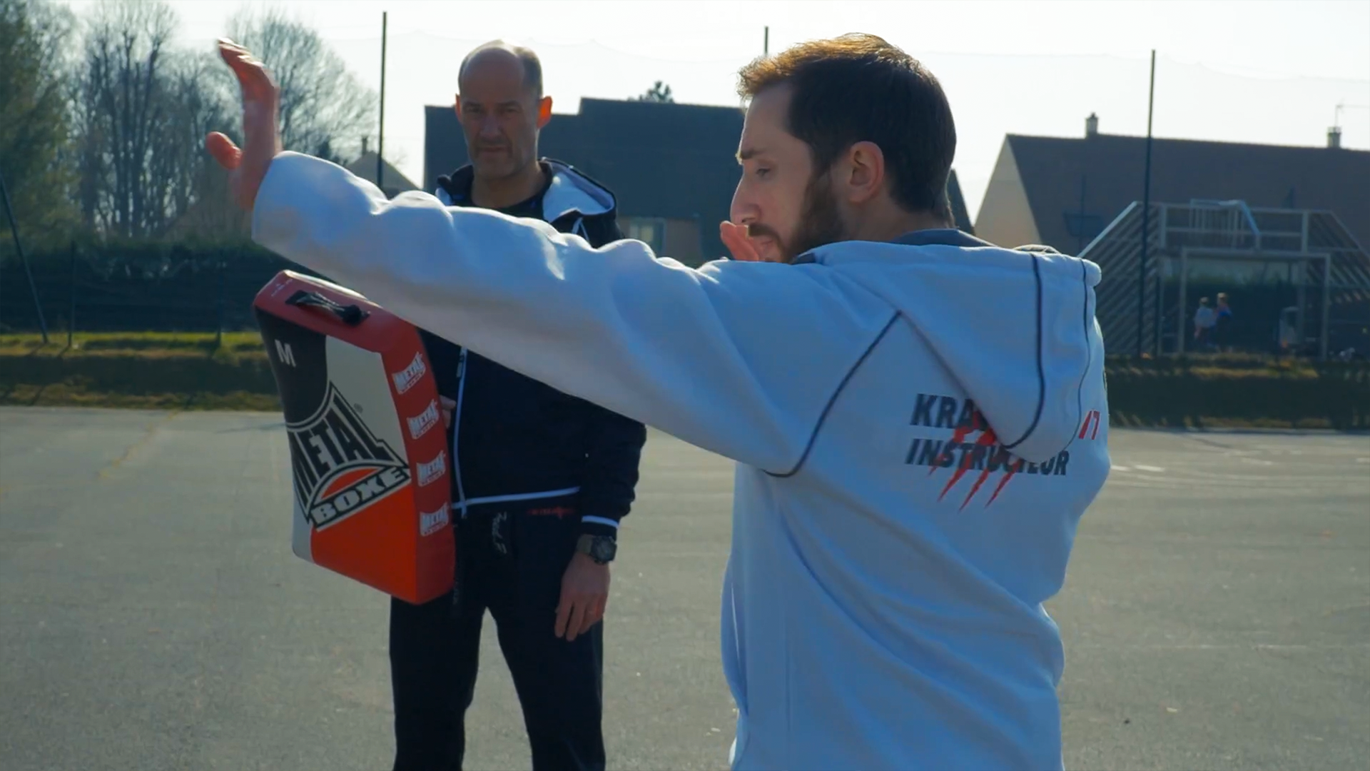 Un instructeurs de krav maga fait une démonstration de posture pour donner un coup de poing défensif. Il est en sweet blanc, devant un autre instructeur qui tien un bouclier de frappe, en extérieur.
