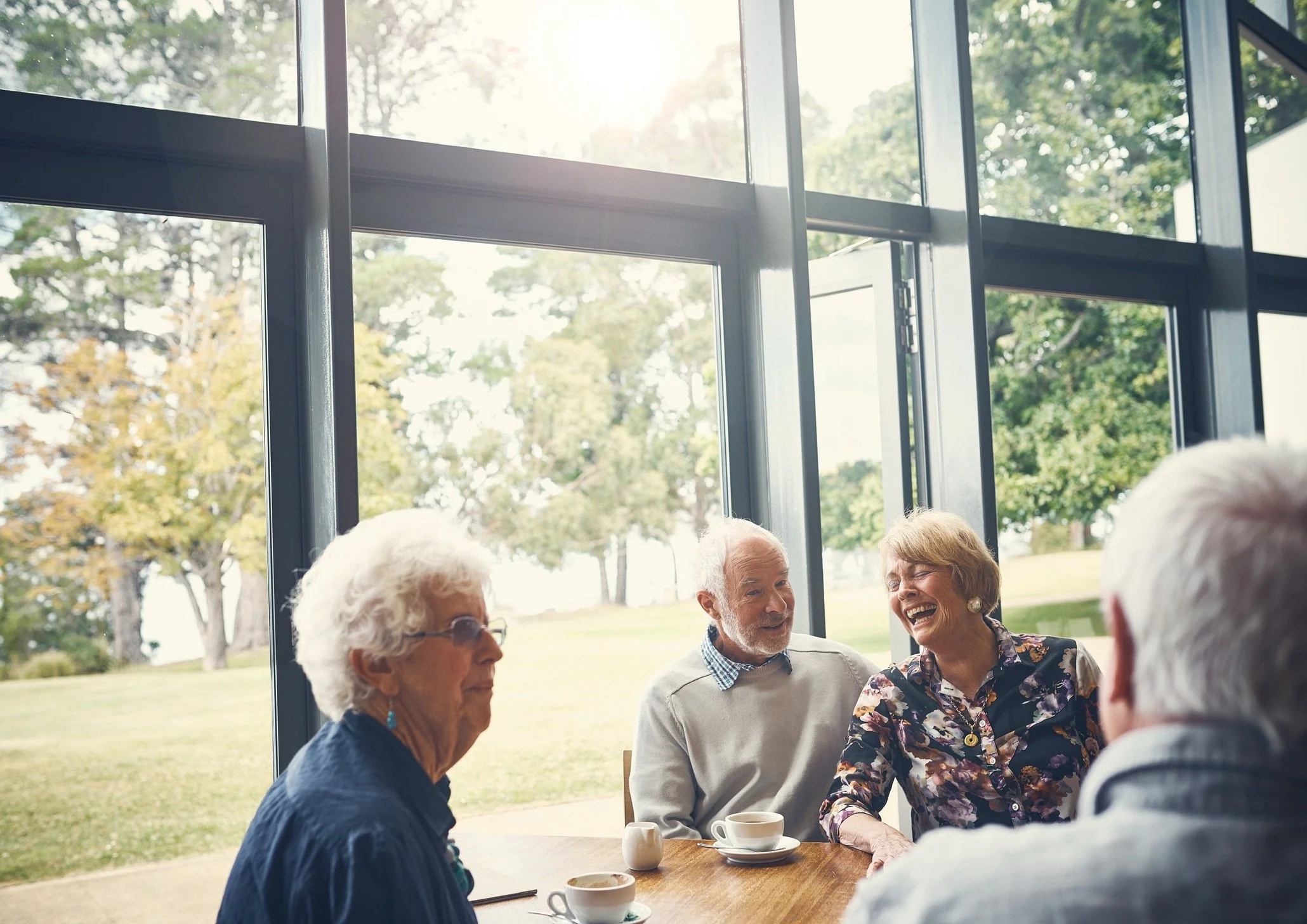Four elderly people sitting at a table having coffee and laughing inside a room with large glass windows showing a green outdoor landscape.