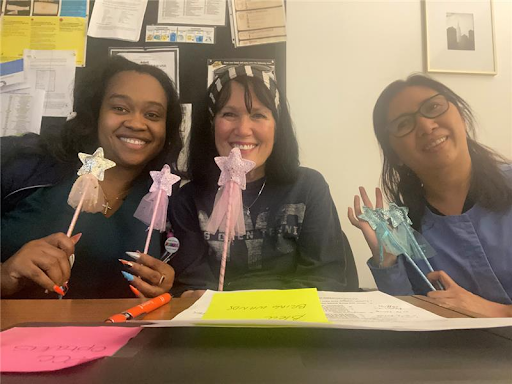 Three women smiling at a table holding star-shaped fairy wands. There are papers, a yellow note, and an orange pen on the table.